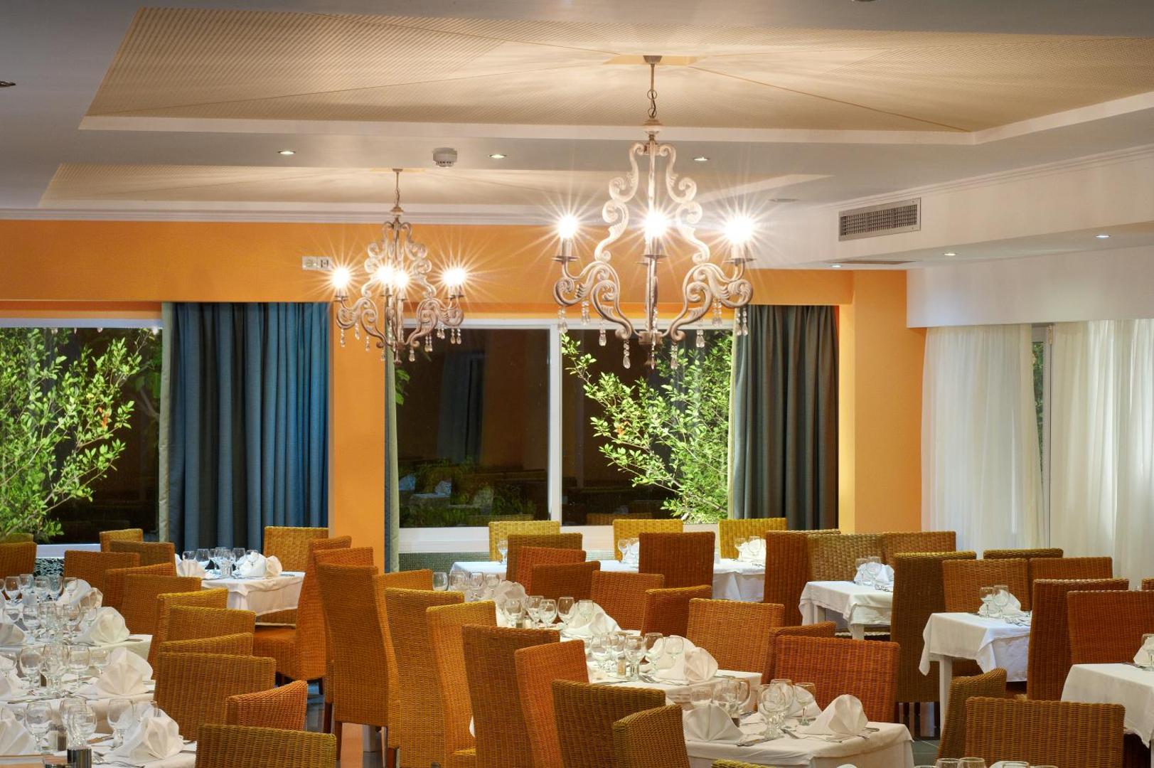 Portes Beach Hotel galvenais restorāns Halkidiki ar klātiem galdiem, pītiem krēsliem un greznām lustrām siltos oranžos toņos