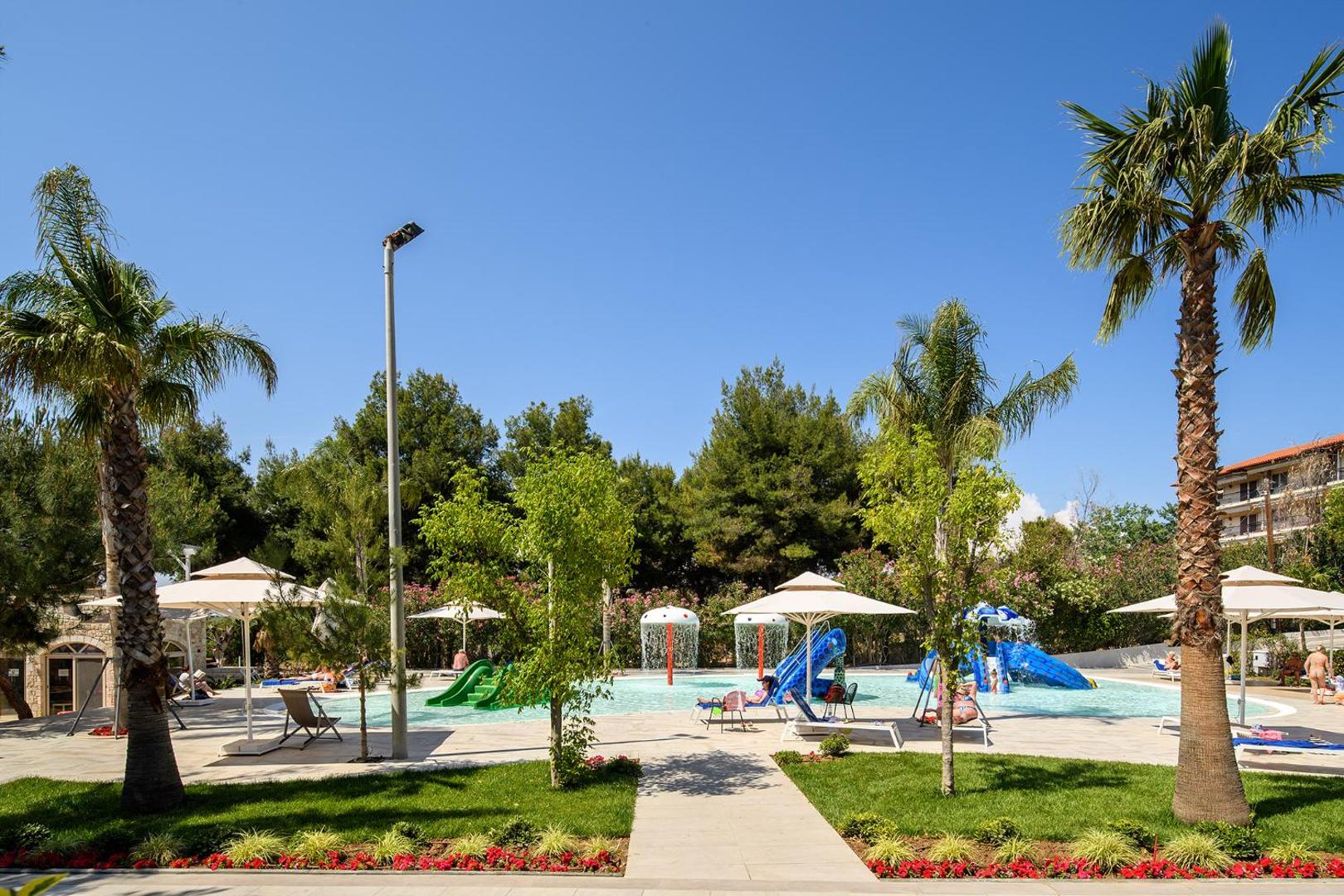 Portes Beach Hotel viesnīcas teritorija Halkidiki ar bērnu baseinu, zaļojošiem zālieniem, palmām un saulessargiem zem skaidrām debesīm