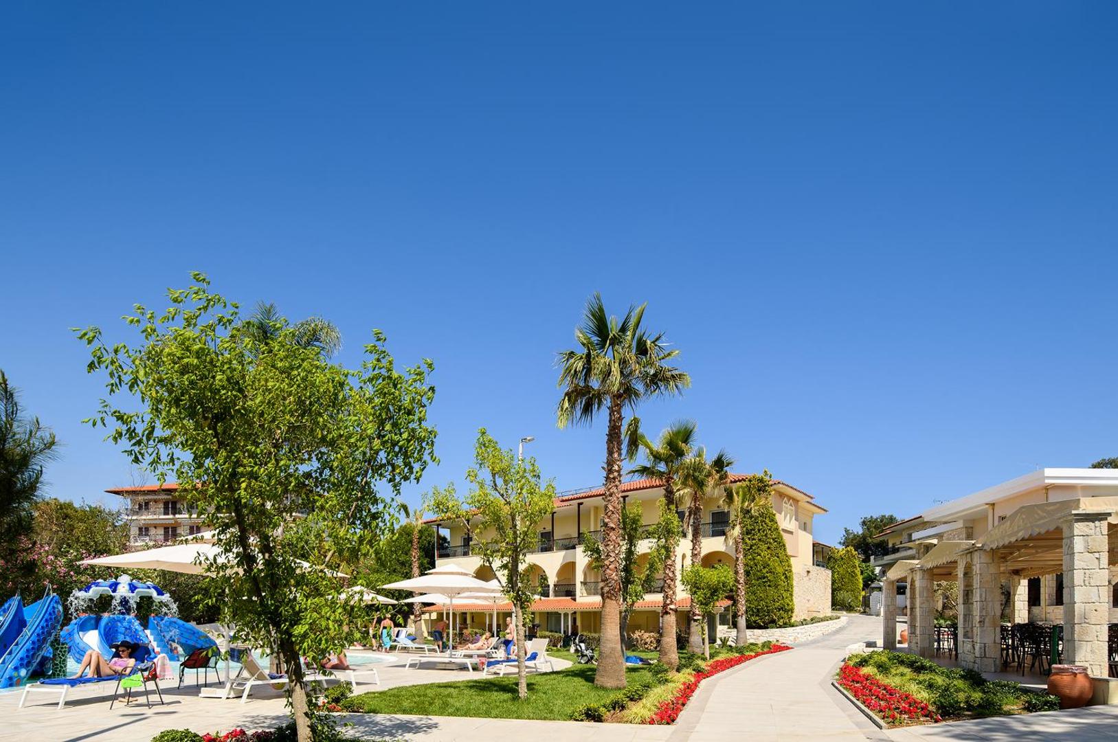 Portes Beach Hotel teritorija ar palmām, ziedošām puķu dobēm un bērnu baseinu ar slidkalniņiem saulainā dienā Halkidiki