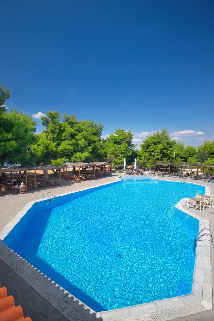 Portes Beach Hotel lielais āra peldbaseins ar zilu ūdeni, ko ieskauj priežu mežs un atpūtas zonas ar saulessargiem