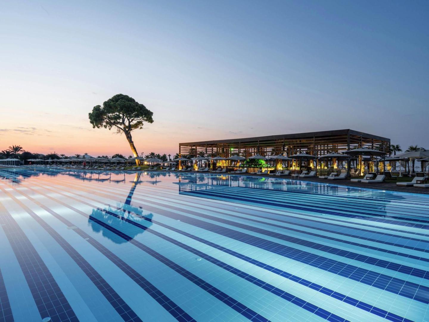 Rixos Premium Belek milzīgs āra baseins saulrieta laikā ar izgaismotu bāru, sauļošanās krēsliem un priežu siluetu pie horizonta