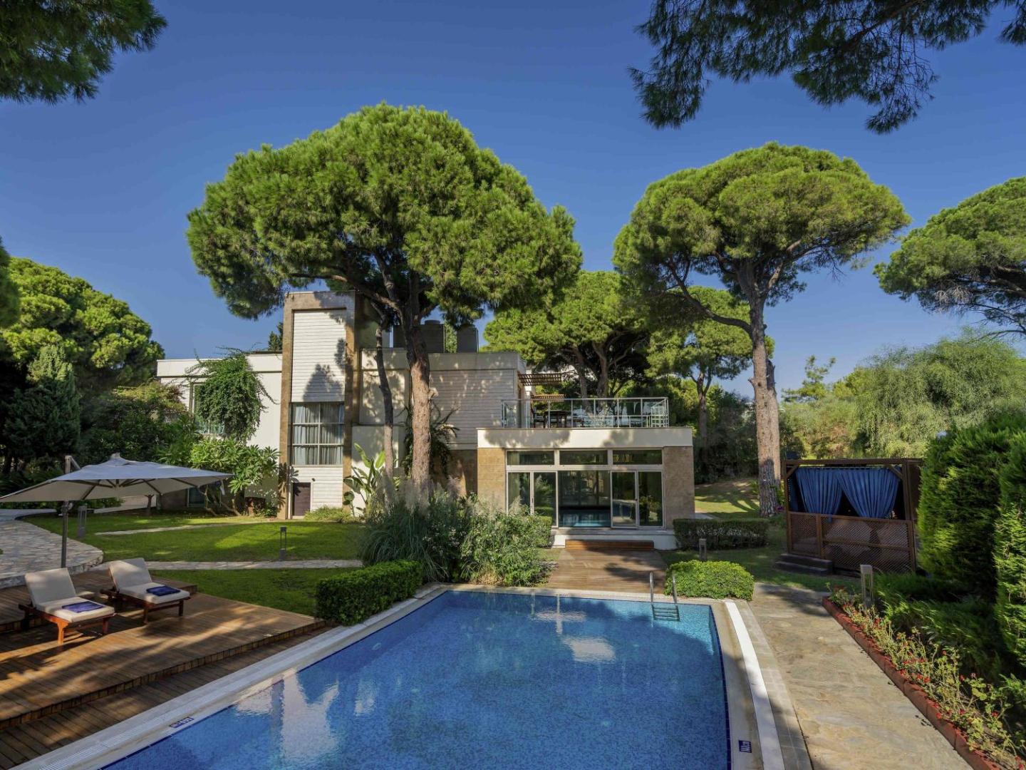Rixos Premium Belek luksusa bungalo ar privātu baseinu, ko ieskauj kupli skuju koki un labiekārtots dārzs ar sauļošanās krēsliem