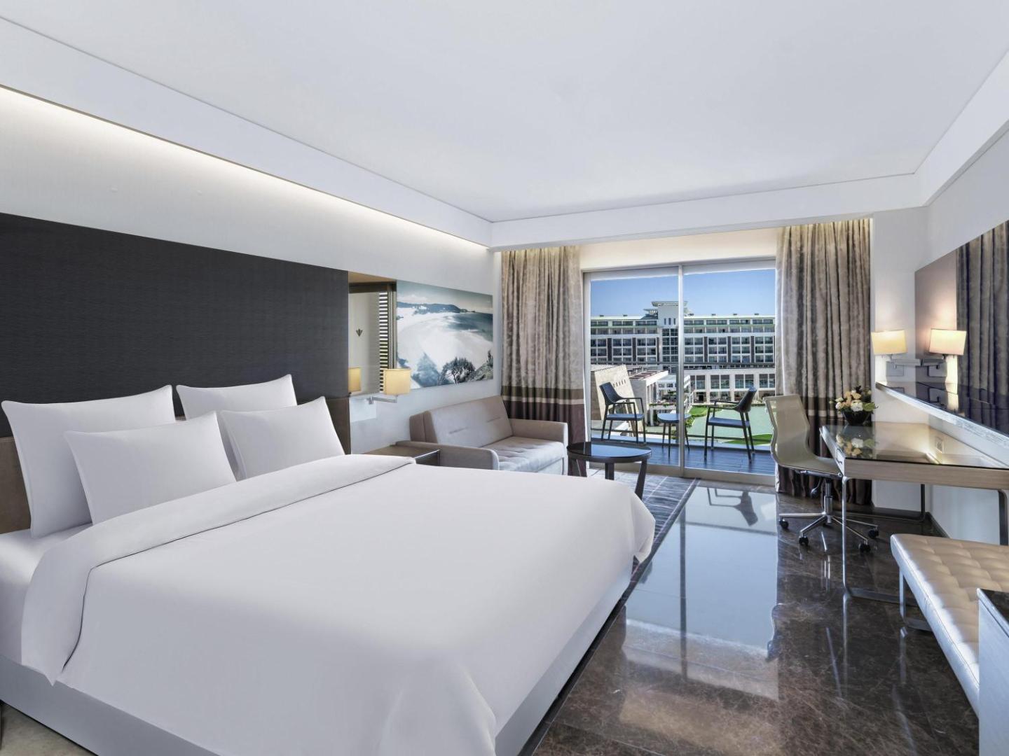 Rixos Premium Belek plašs numurs ar atpūtas zonu, dīvānu un stiklotu sienu, kas ved uz balkonu ar skatu uz viesnīcas teritoriju