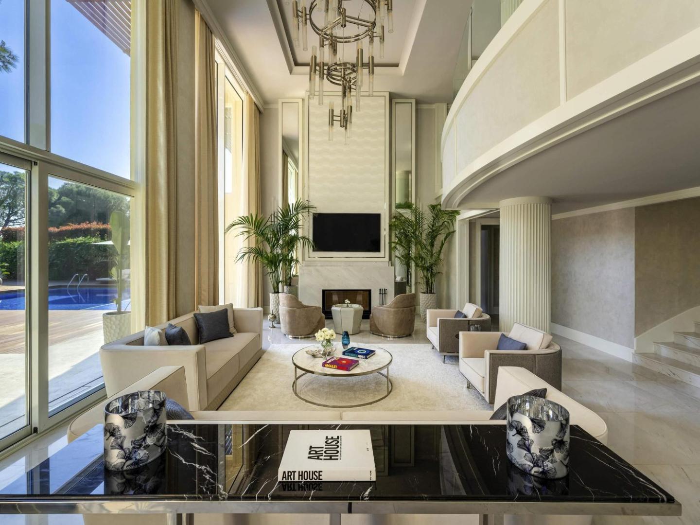 Rixos Premium Belek grezna atpūtas zona ar augstiem griestiem, kamīnu, mīkstajiem krēsliem un skatu uz āra baseinu caur lieliem logiem