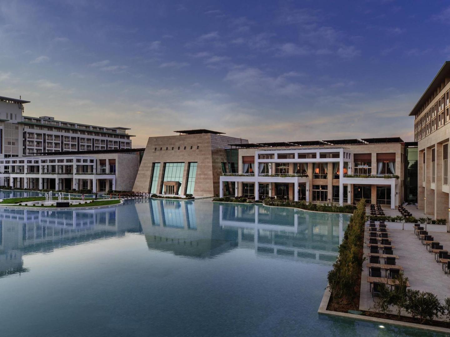 Rixos Premium Belek galvenā ēka un terases pie liela dekoratīvā baseina saulrieta laikā Belekā