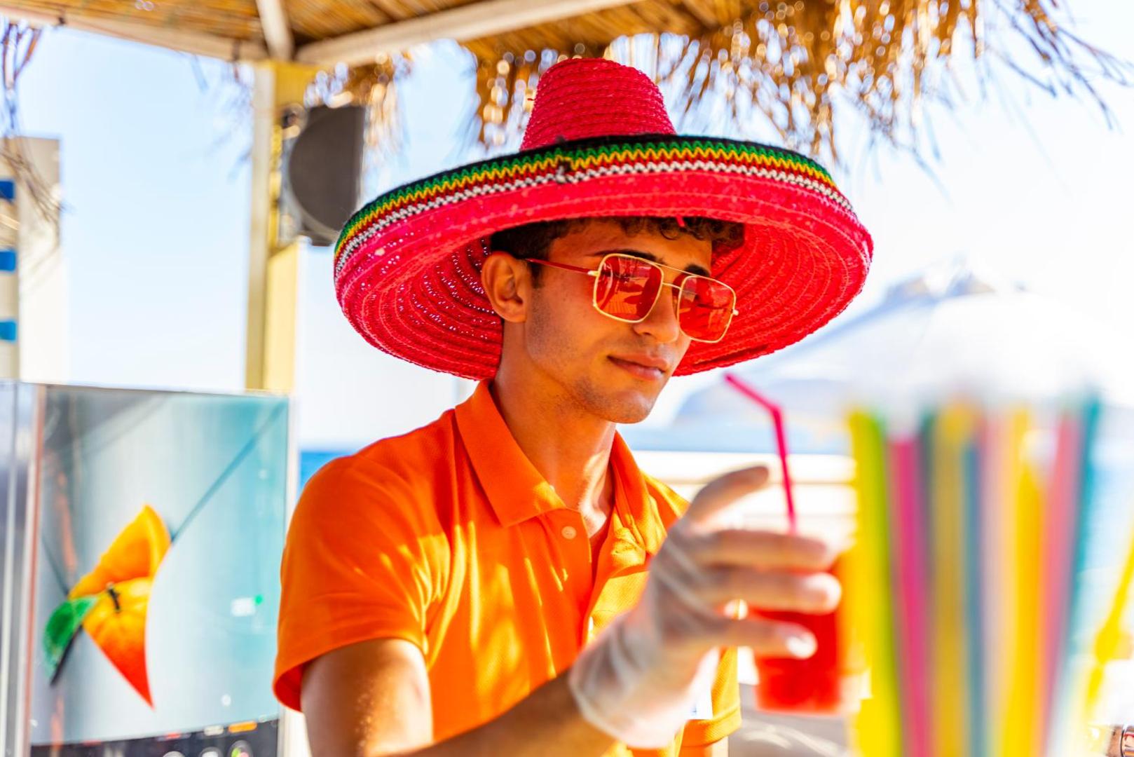 Ulusoy Kemer Holiday Club bārmenis košā cepurē pasniedz atvēsinošu dzērienu pludmales bārā Kemeras piekrastē