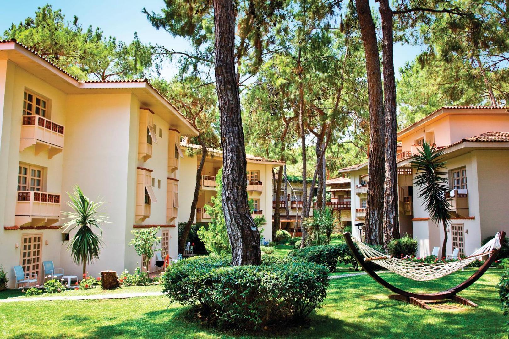 Ulusoy Kemer Holiday Club teritorija ar šūpuļtīklu zālienā un gaišām viesnīcas ēkām, ko ieskauj Vidusjūras reģiona priedes
