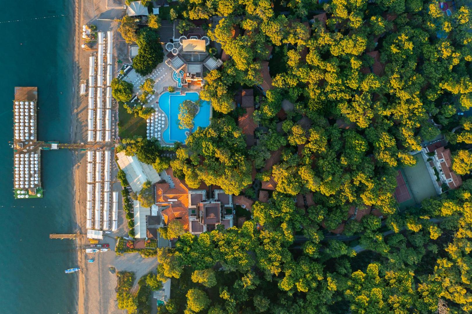 Ulusoy Kemer Holiday Club teritorijas skats no augšas, parādot baseinu kompleksu, tenisa kortus un blīvu zaļo zonu pie jūras
