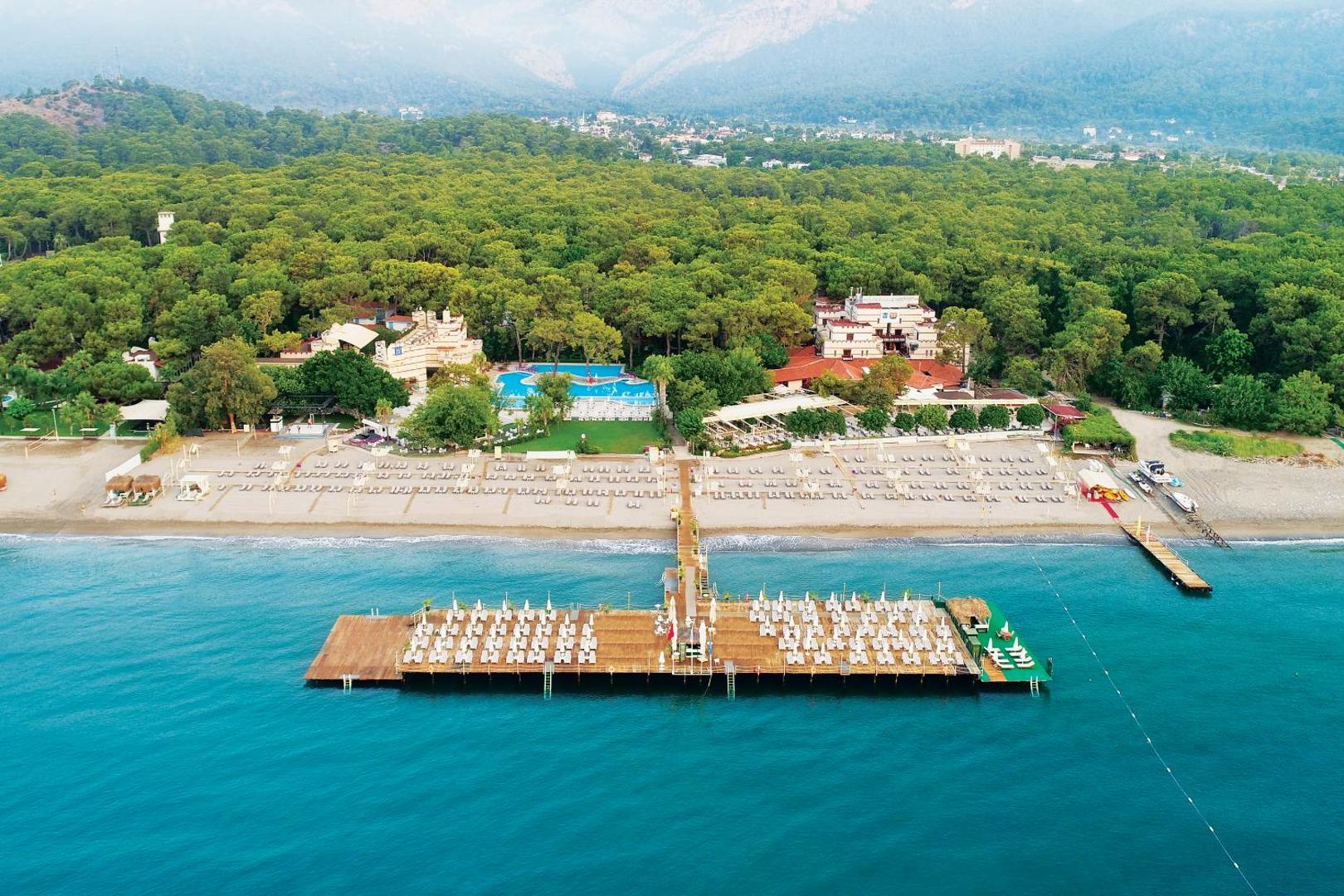 Ulusoy Kemer Holiday Club panorāmas skats no putna lidojuma uz pludmali, koka piestātni ar sauļošanās krēsliem un apkārtējo priežu mežu