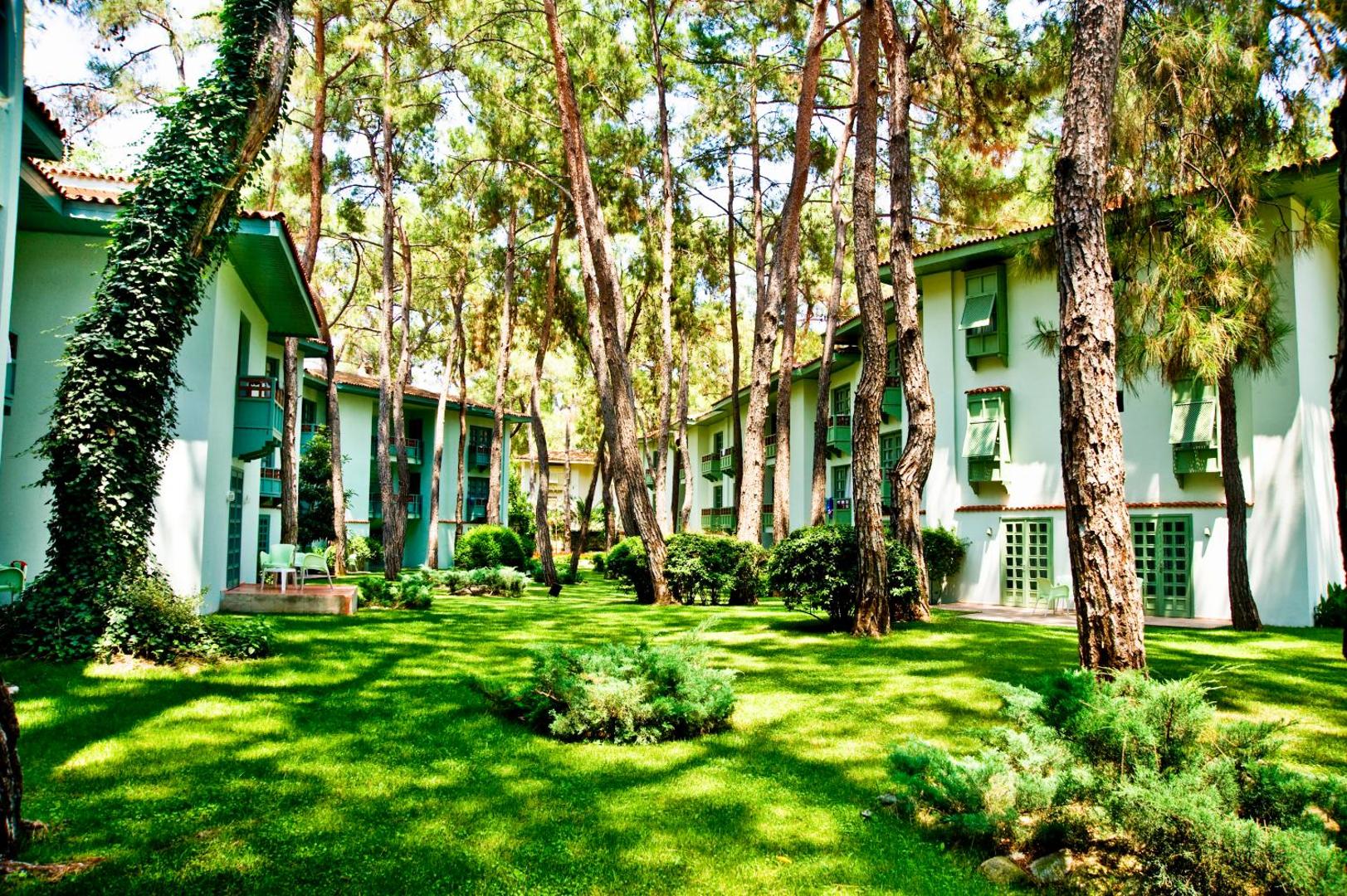 Ulusoy Kemer Holiday Club viesnīcas numuru ēkas ar zaļiem slēģiem, kas izvietotas starp augstām priedēm un koptu mauriņu