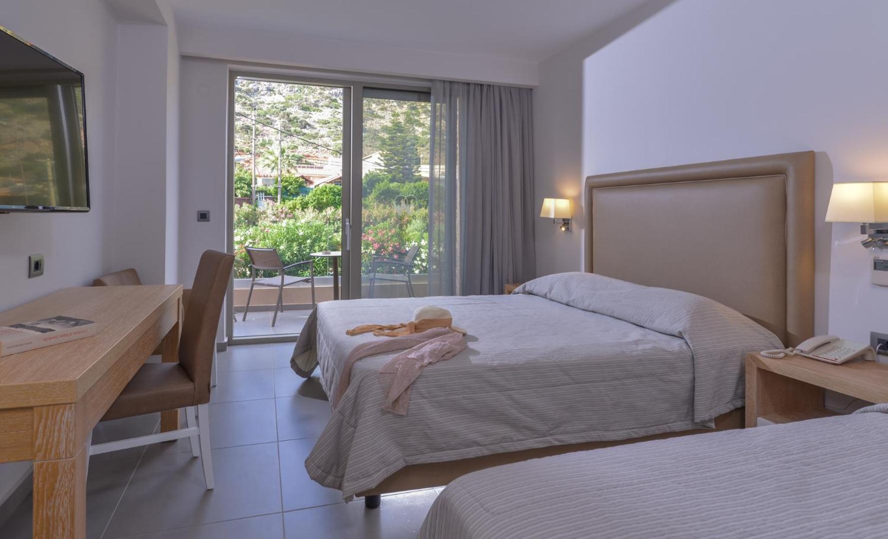 Gaišs viesnīcas numurs ar divguļamo gultu un skatu uz dārzu caur balkonu.