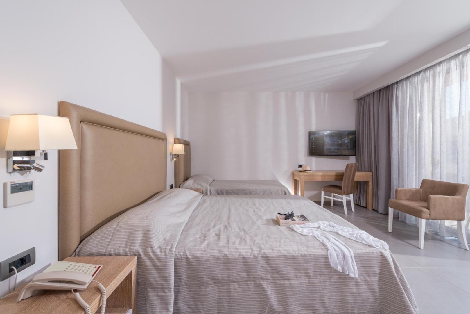 Gaišs numurs ar divām gultām, darba galdu, televizoru un atpūtas krēslu.