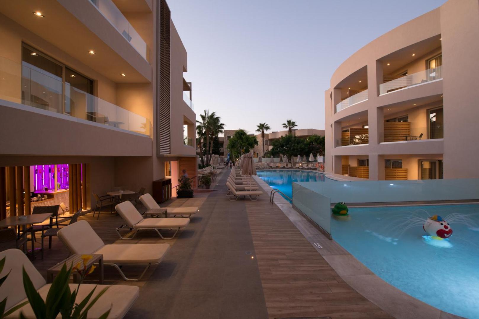 Viesnīcas pagalms vakarā ar baseinu, bērnu baseinu un sauļošanās krēsliem.