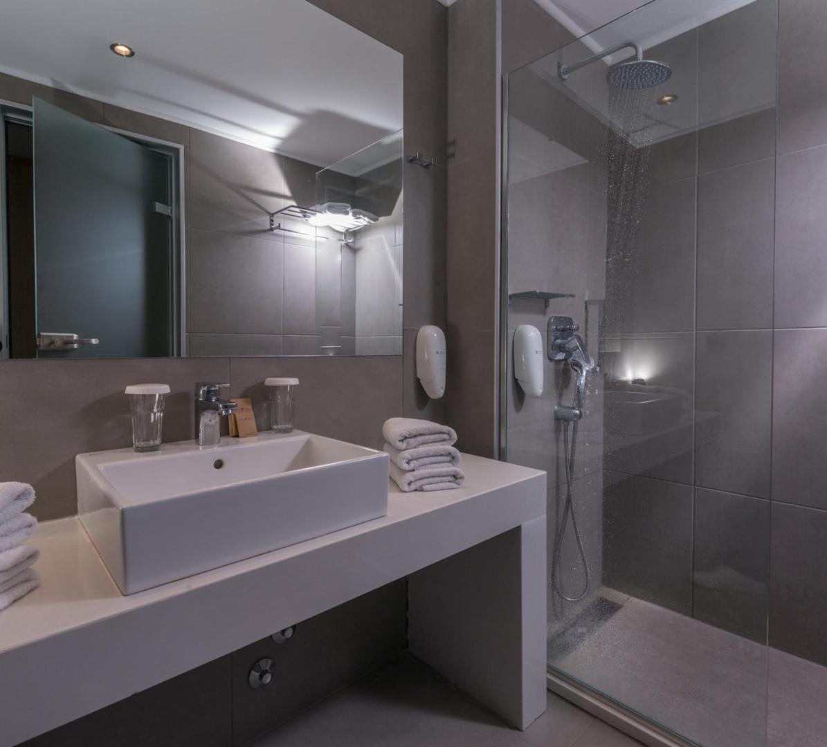 Moderna viesnīcas vannas istaba ar pelēkām flīzēm, baltu izlietni un stikla dušas kabīni.