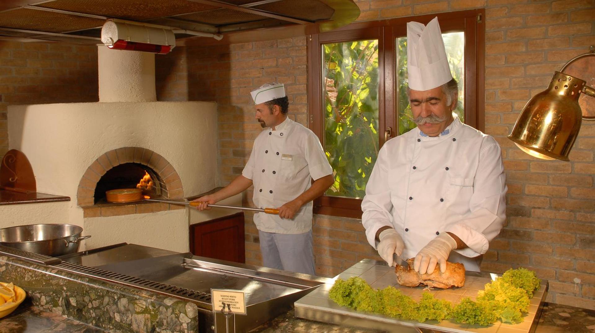 Divi pavāri gatavo ēdienu pie tradicionālas mūra krāsns un griež gaļu.