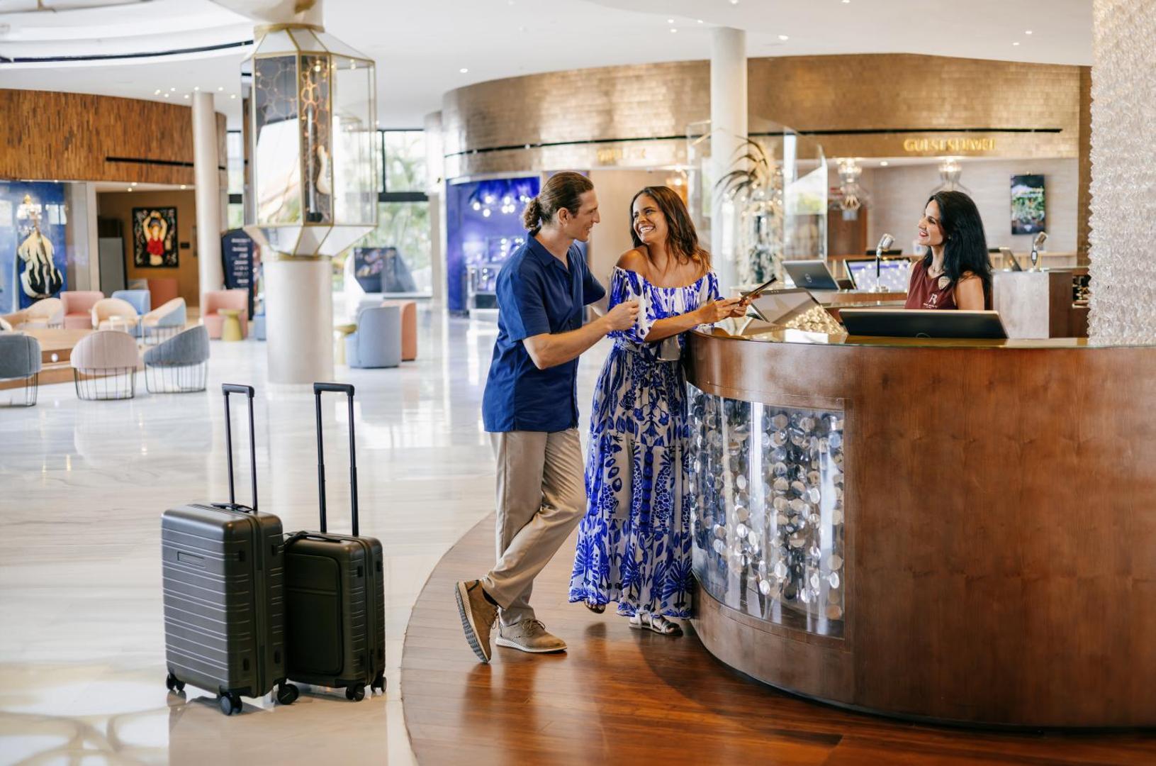 Viesi reģistrējas Hard Rock Hotel Tenerife pie koka apdares recepcijas galda, fonā redzami koferi un moderns vestibila interjers.