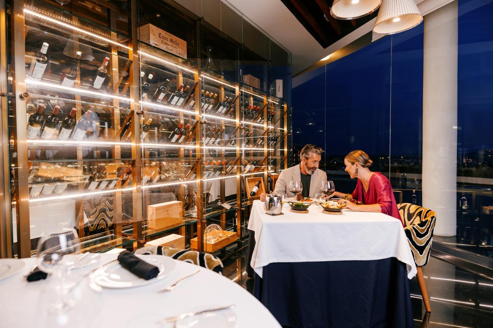 Hard Rock Hotel Tenerife restorāns ar plašu vīna skapi un pāri, kas bauda vakariņas pie eleganti klāta galda Tenerifē.