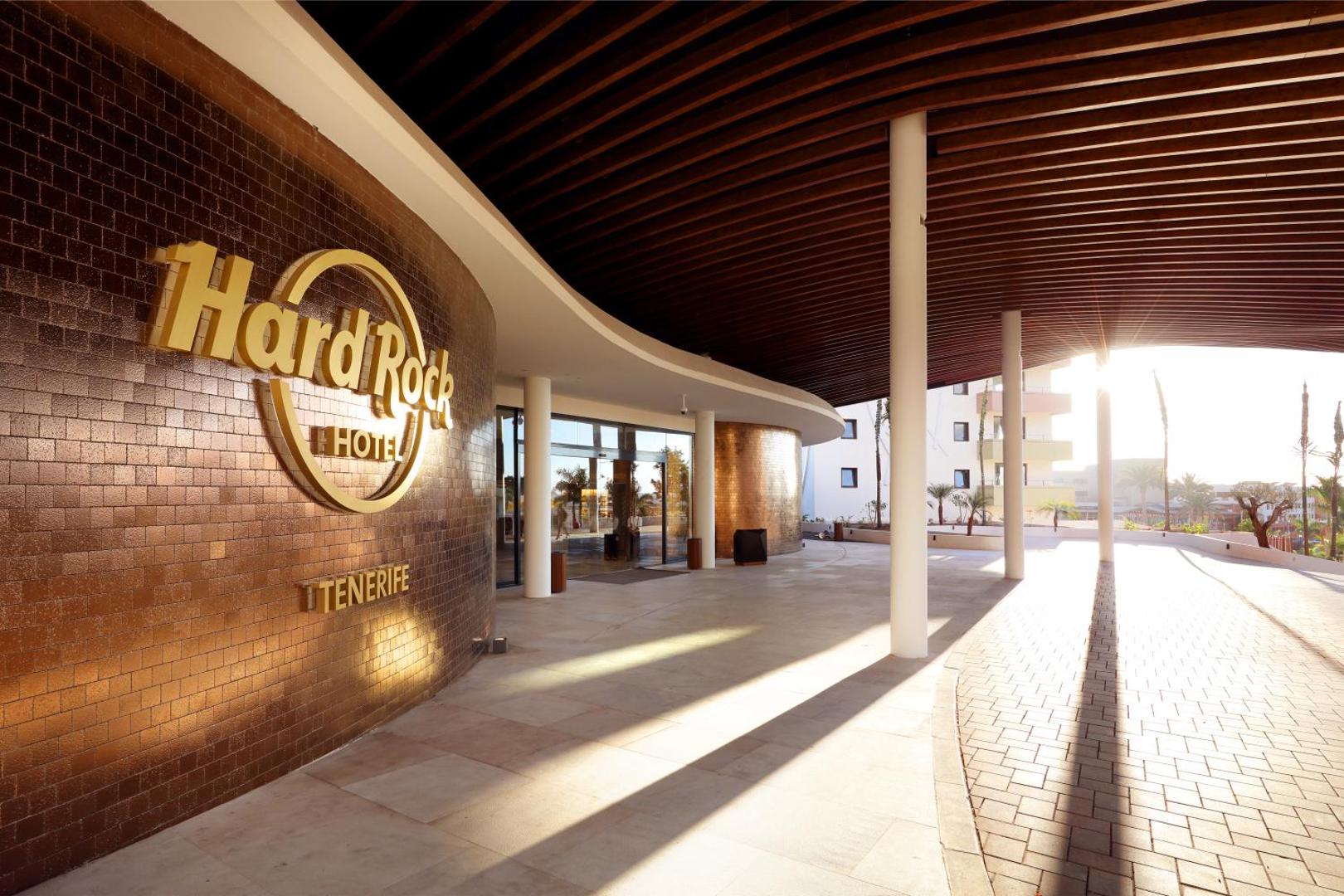 Hard Rock Hotel Tenerife ieeja ar zeltītu logo uz brūnu flīžu sienas un saules apspīdētu kolonādi piekrastē.