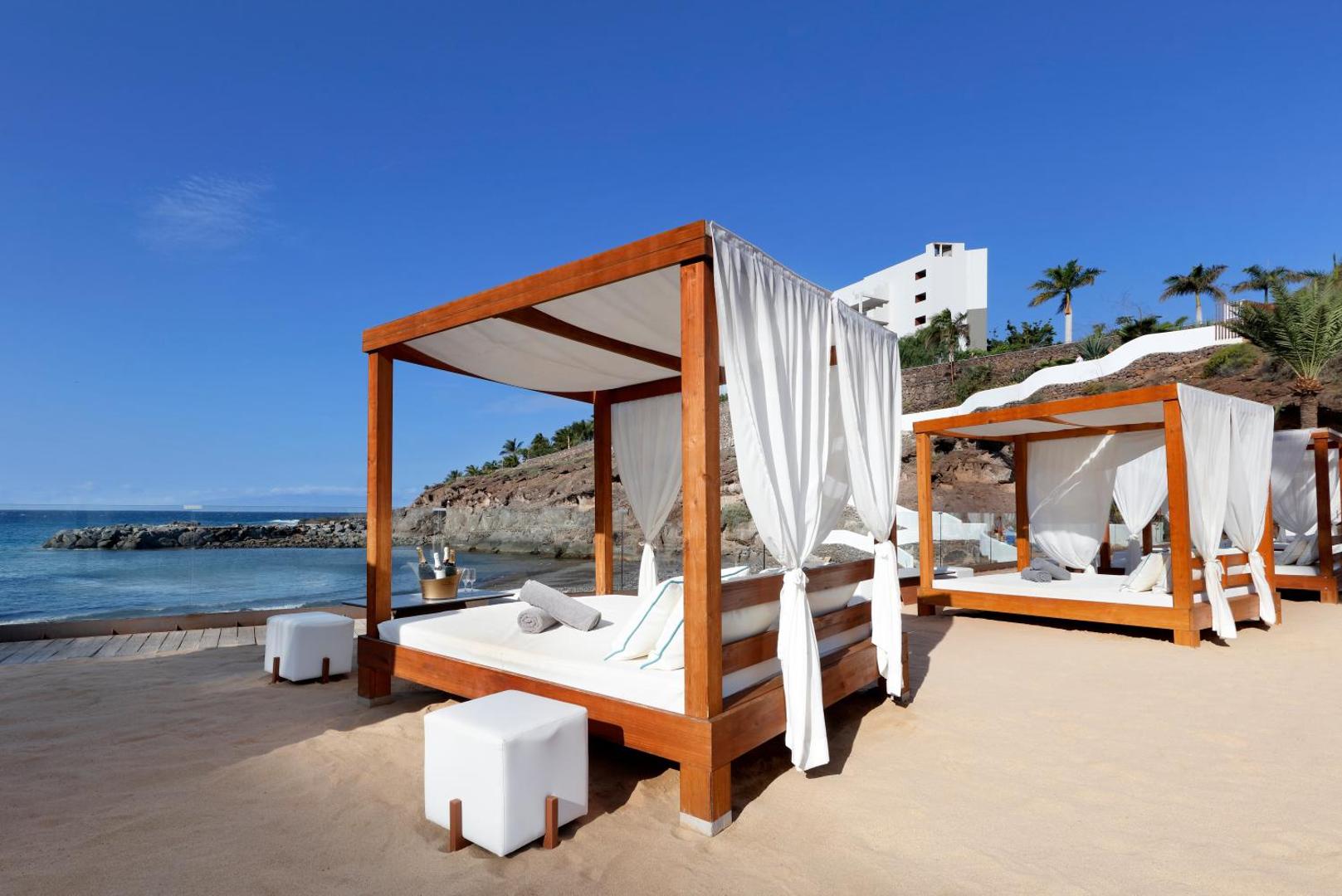 Hard Rock Hotel Tenerife atpūtas zona pludmalē ar koka bungalo tipa gultām un baltiem aizkariem pie okeāna krasta.