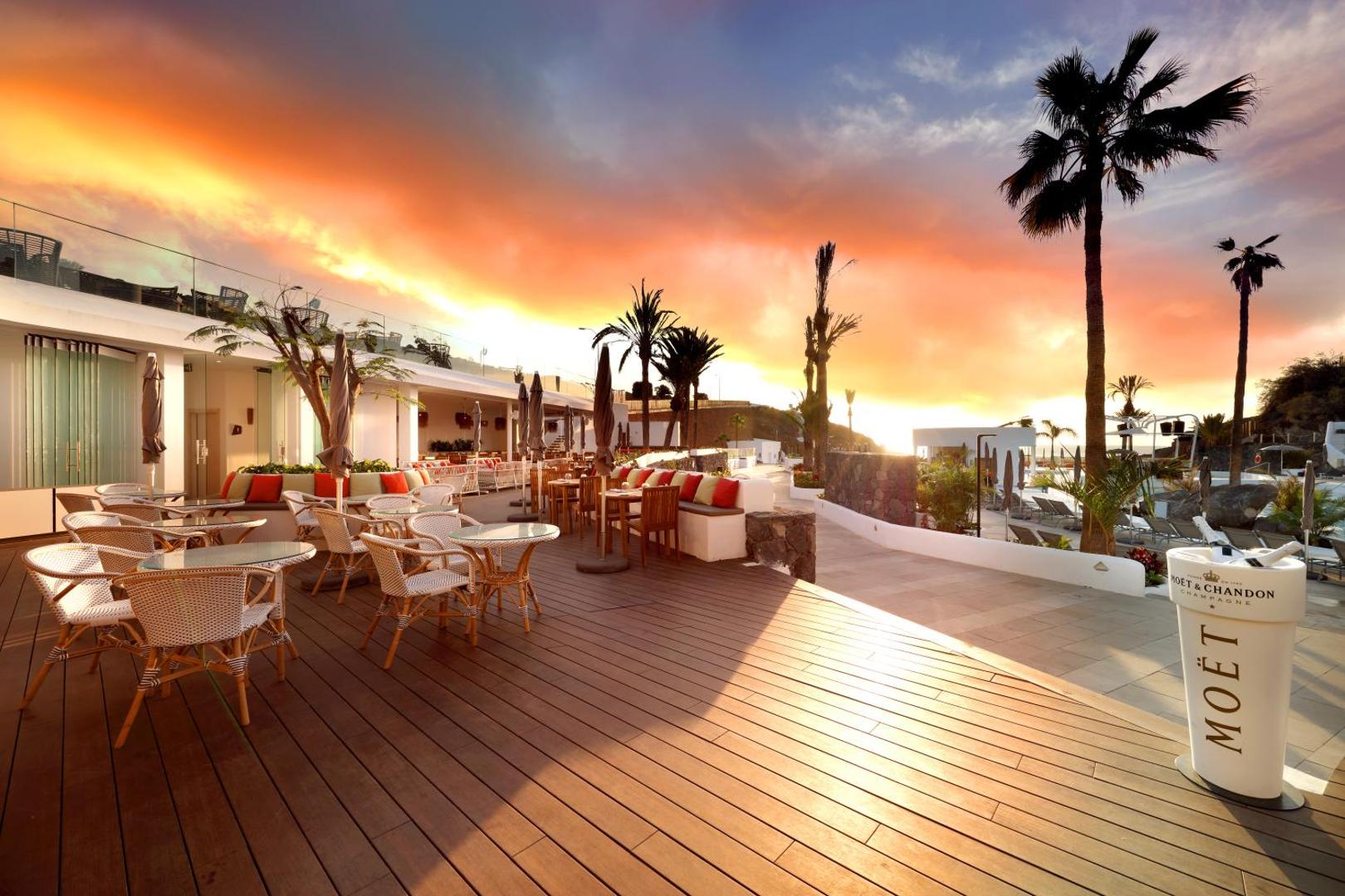 Hard Rock Hotel Tenerife āra terase ar galdiņiem un dīvāniem krāšņa saulrieta laikā pie okeāna piekrastes