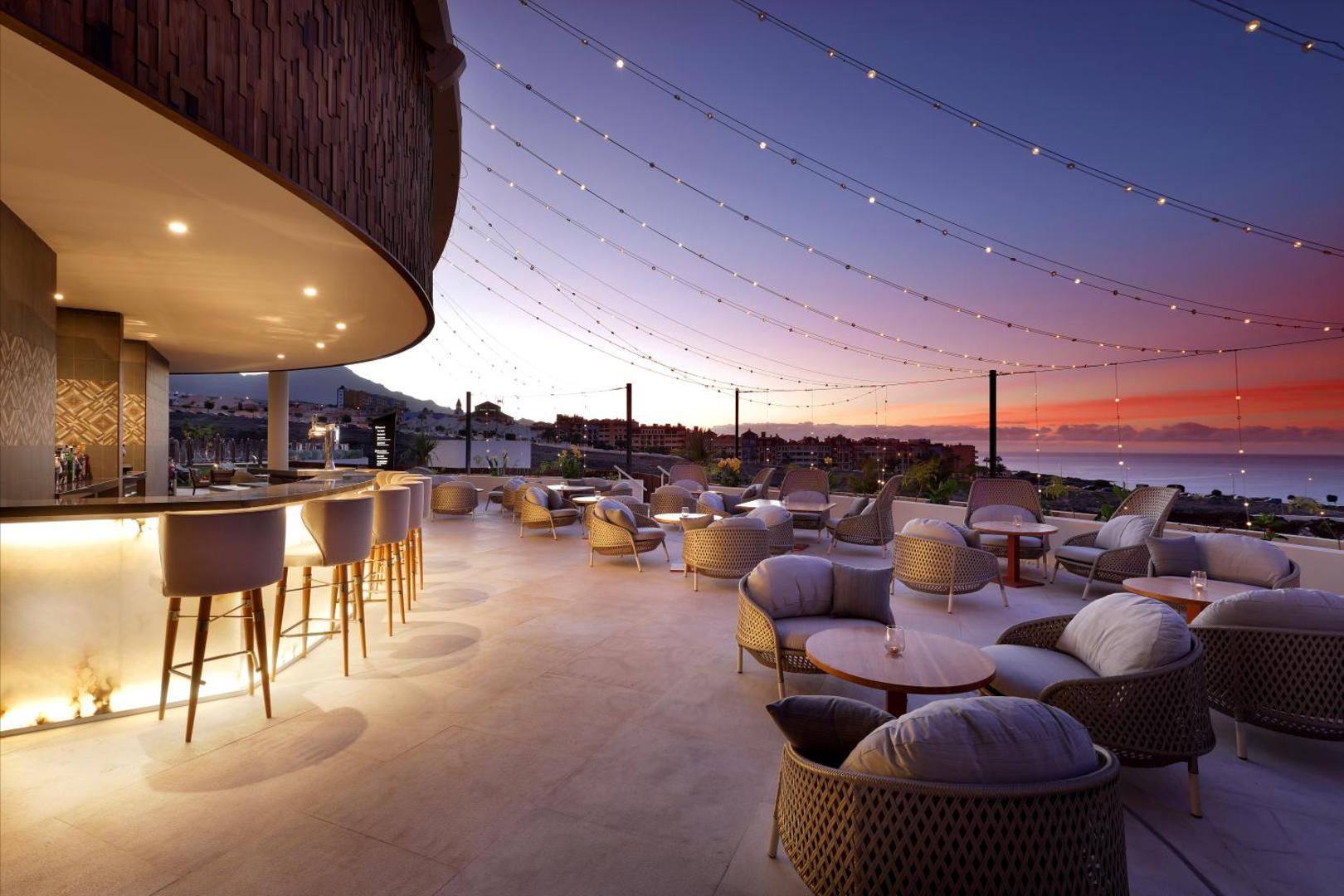 Hard Rock Hotel Tenerife jumta terases bārs saulrietā ar ērtiem krēsliem, dekoratīvām gaismām un skatu uz okeānu Tenerifē.
