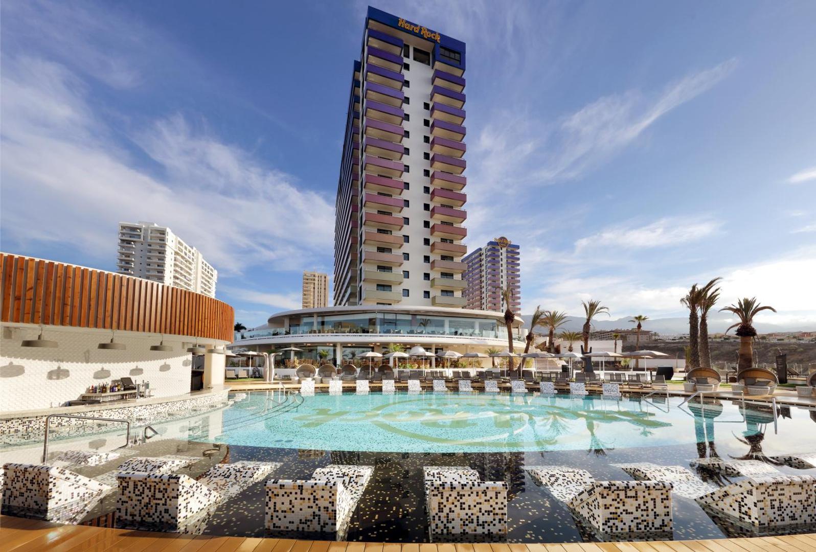 Hard Rock Hotel Tenerife āra baseins ar atpūtas krēsliem ūdenī un skatu uz viesnīcas augstceltni Tenerifes dienvidu piekrastē.