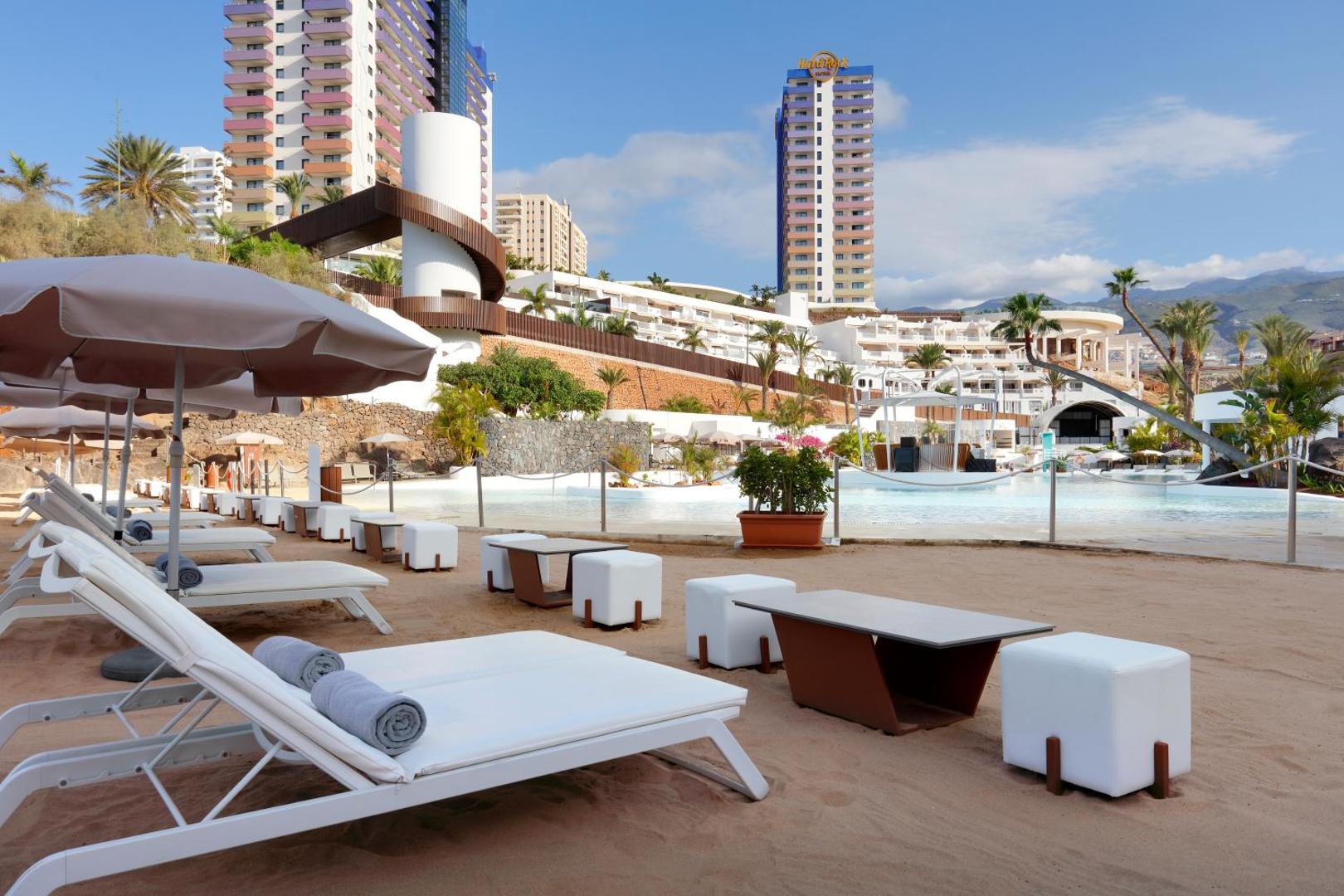Hard Rock Hotel Tenerife kopskats ar baseina zonu, sauļošanās krēsliem un augstceltņu korpusiem uz kalnu un palmu fona.