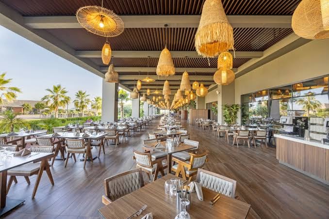 Jacaranda Luxury Resort āra terases restorāns ar modernām mēbelēm un stilīgiem pītiem gaismekļiem saulainā dienā
