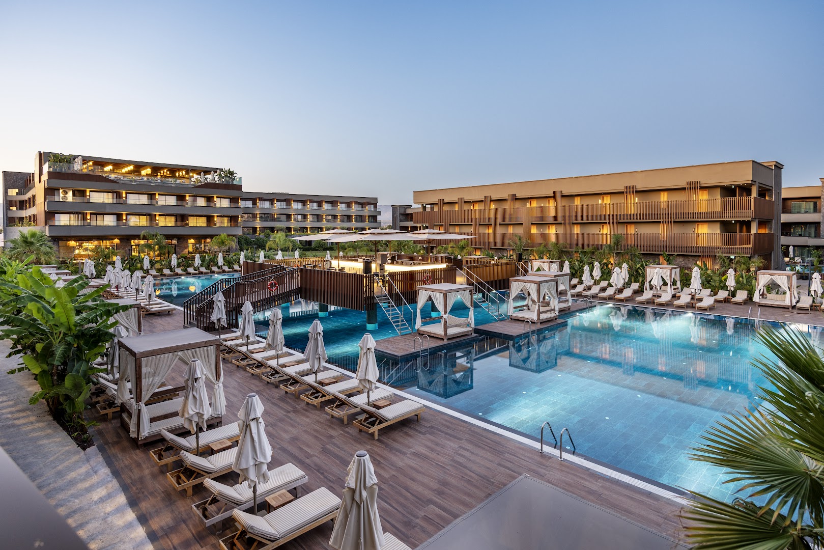 Jacaranda Luxury Resort lielais āra baseins vakarā ar izgaismotām atpūtas nojumēm, sauļošanās krēsliem un viesnīcas korpusiem fonā