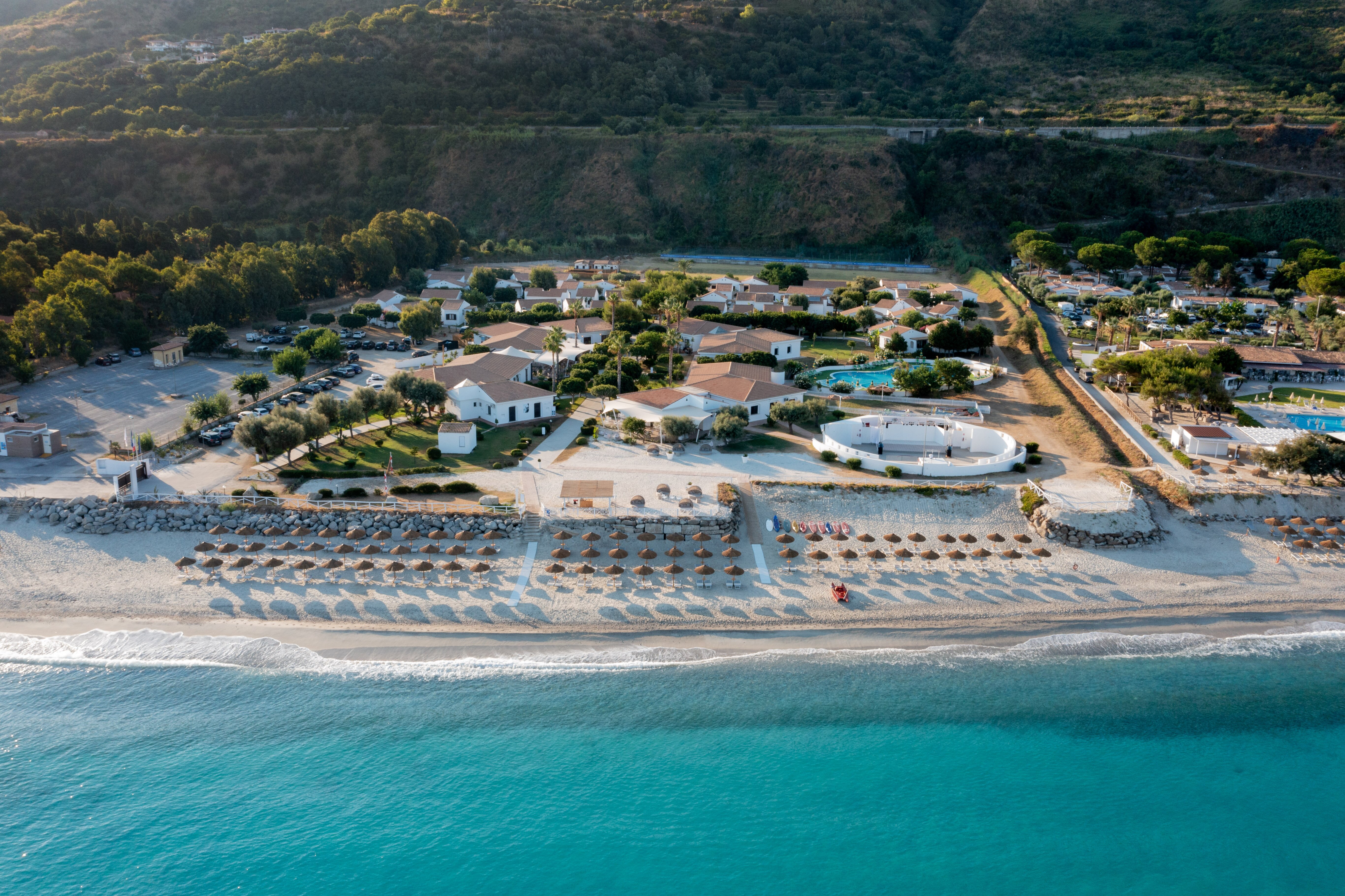 Pludmales kūrorts ar saulessargiem un viesnīcām, kas atrodas pie jūras. Aiz pludmales redzama zaļa daba un kalni.