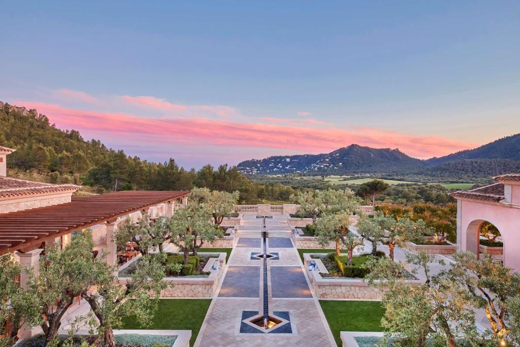 Eleganta ainava ar ainavu, kurā redzams dārzs ar olīvkokiem un akmens celiņiem, apgaismota saulrieta gaismā. Fons veido kalni un zaļumi, radot mierīgu un idillisku atmosfēru.