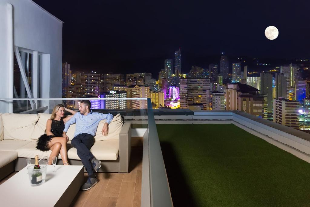 Pāris atpūšas uz jumta dīvāna, skatoties uz pilsētas apgaismotajiem debesskrāpjiem nakts laikā, kad gaisā ir pilna mēness.