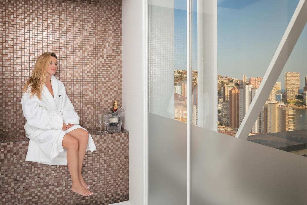 Sieviete baltā halātā atpūšas uz mozaīkas sienas, blakus ir šampanieša pudele un glāzes, aiz loga paveras pilsētas panorāma.