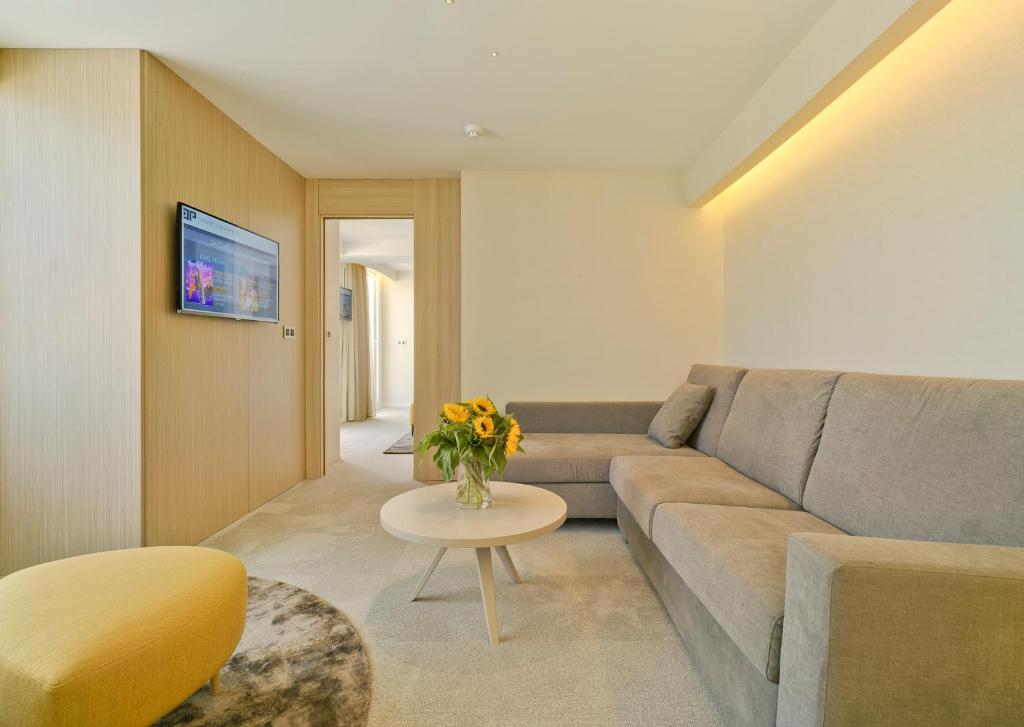 Mūsdienīgs viesistabas interjers ar pelēku dīvānu, apaļu galdiņu un saulainu ziedu pušķi, kā arī televizoru uz sienas. Telpa ir gaiša un gaumīga, radot mājīgu atmosfēru.