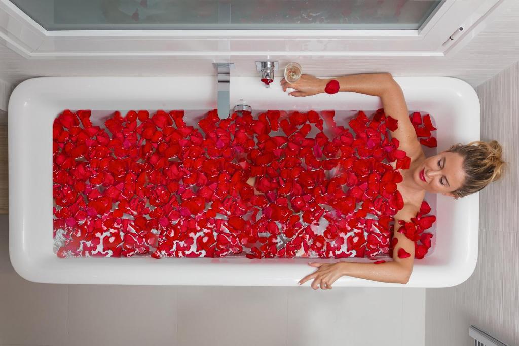 Sieviete atpūšas vannā, kas piepildīta ar sarkaniem rožu ziedlapiņām, tur rokā glāzi dzēriena. Vanna ir moderna un gaiša, radot relaksējošu atmosfēru.