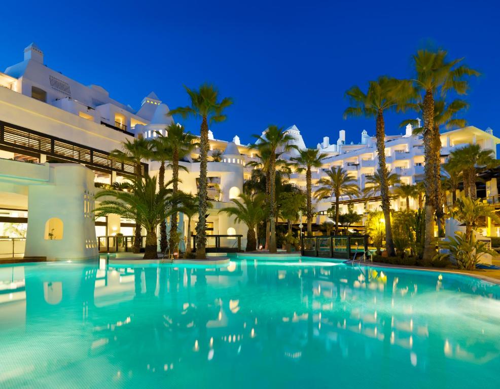 Viesnīcas komplekss ar baseinu, palmām un apgaismotu arhitektūru, kas izgaismota vakara debesīs. Ideāla vieta relaksācijai un atpūtai.