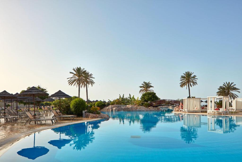 Atlantica Imperial Resort baseins ar palmām un sauļošanās krēsliem piekrastē.