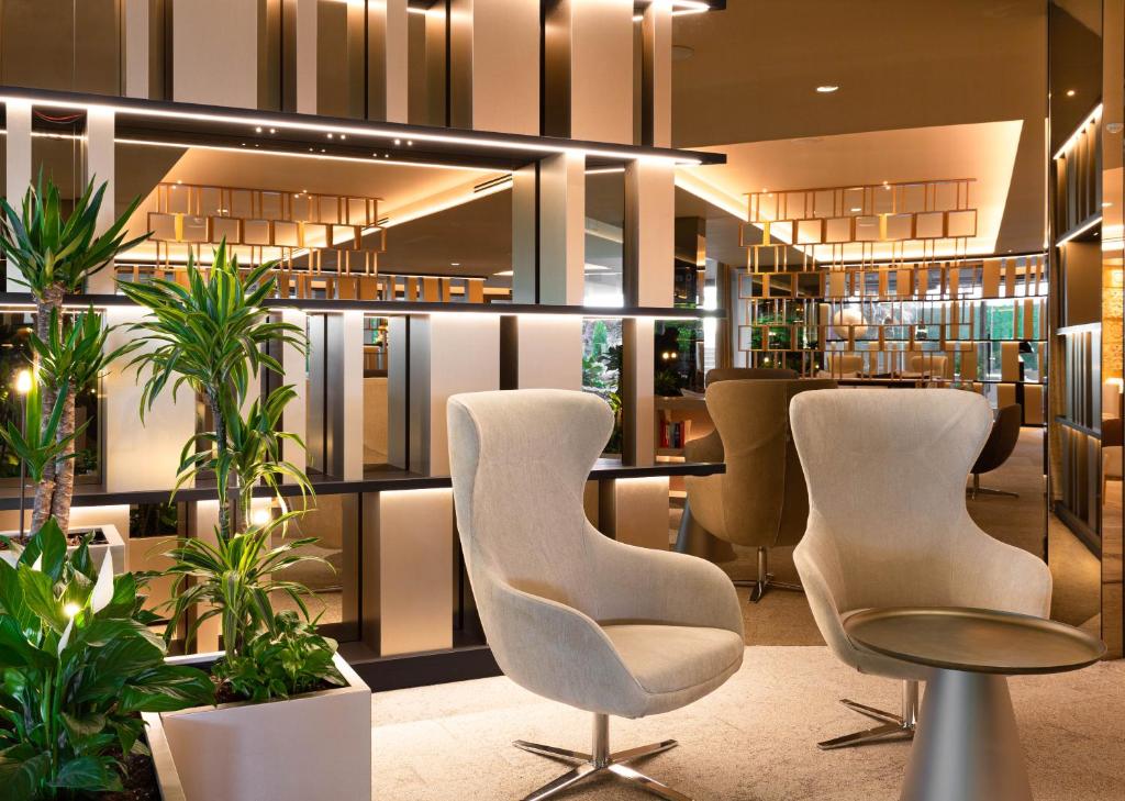 Mūsdienīga interjera dizaina telpa ar ērtām krēsliem un zaļām augiem, radot relaksējošu atmosfēru. Gaismas un spoguļu elementi piešķir telpai eleganci un plašuma sajūtu.