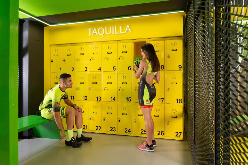 Sporta drēbēs ģērbušies cilvēki stāv blakus dzeltenām skapīšiem sporta zālē, ar zaļu soli un mūsdienīgu interjeru.