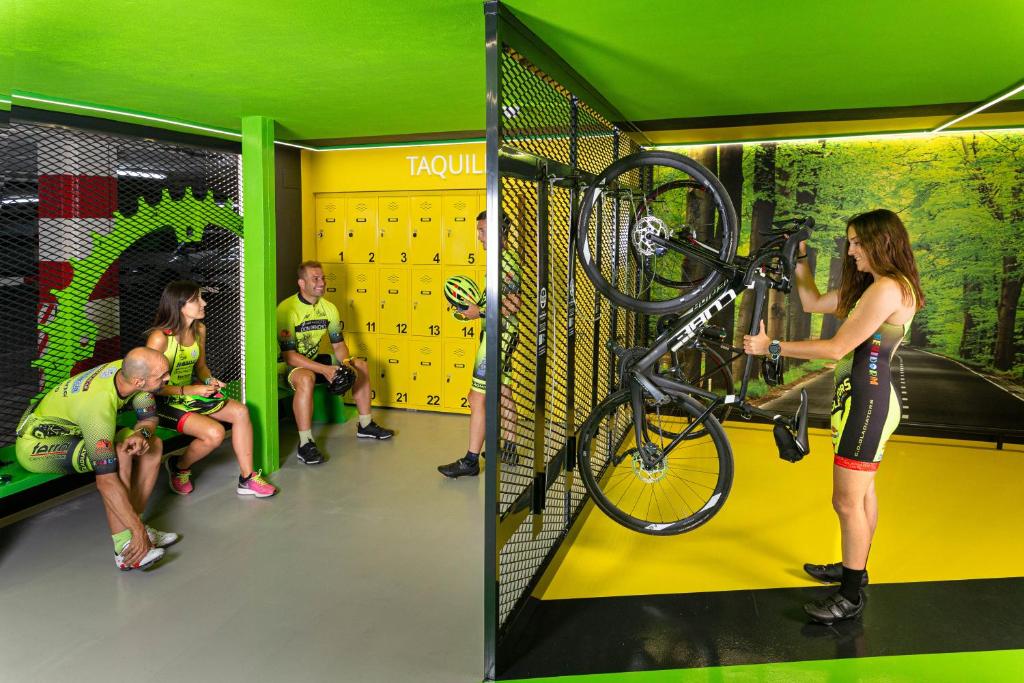 Velosipēdisti atpūšas un sagatavojas braucienam, kamēr kāda sieviete uzglabā savu velosipēdu pie sienas. Telpa ir gaiša ar zaļiem un dzelteniem akcentiem.