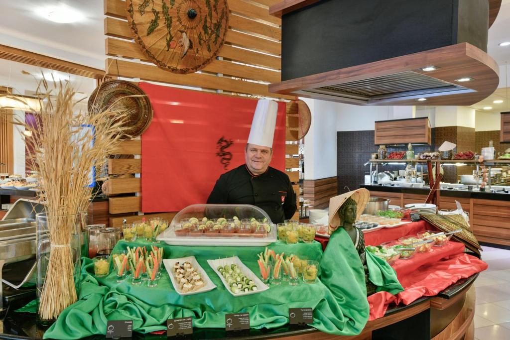 Šefpavārs stāv pie bagātīgi klāta bufetes galda, kurā redzami dažādi ēdieni un dekorācijas, ietverot dārzeņu uzkodas un krāsainus salātus. Fons ir rotāts ar sarkanu audumu un tradicionāliem elementiem.