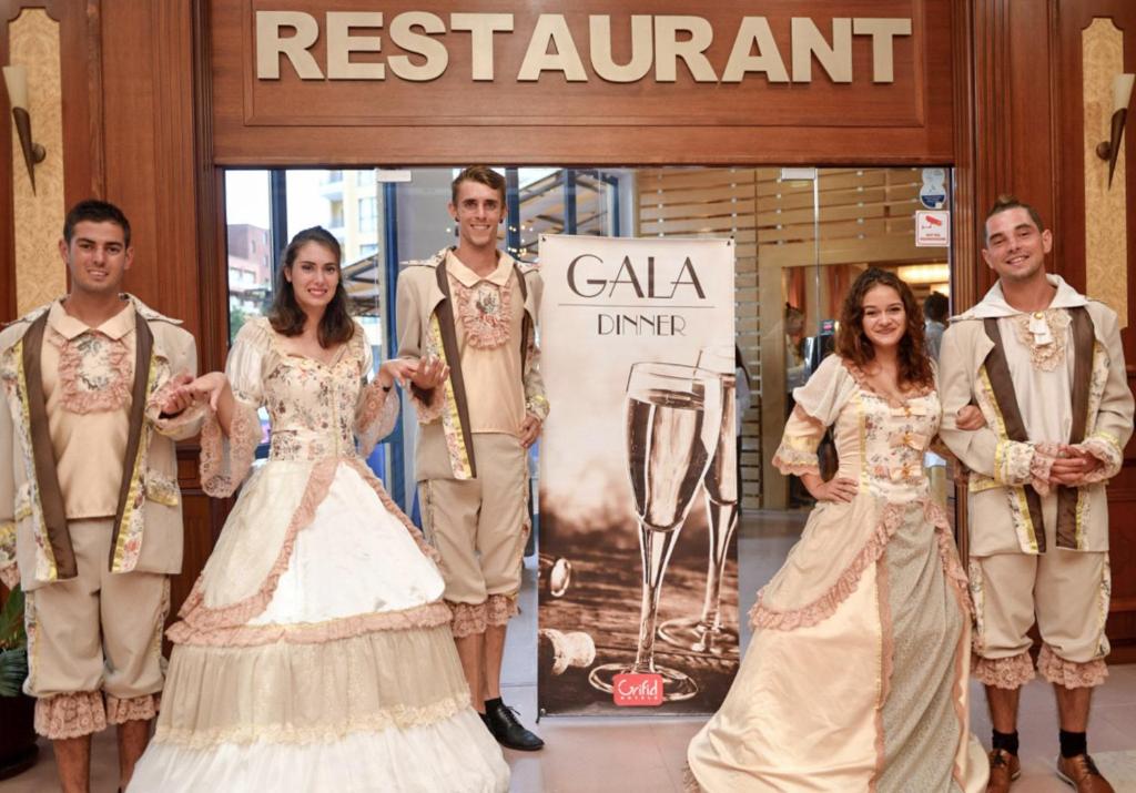 Restorāna ieeja ar viesmīļiem, tērptiem elegantos kostīmos, kas pozē pie plakāta, kas reklamē GALA vakariņas.