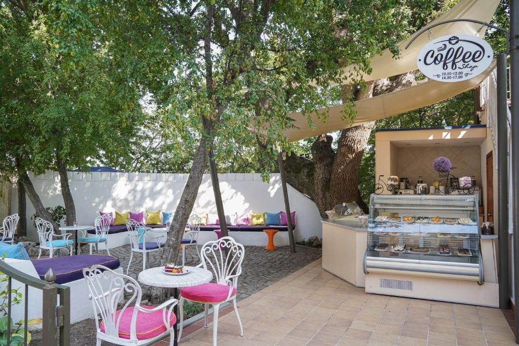 Kafejnīcas āra zona ar krāsainām mēbelēm un gardumiem vitrīnā, ko ieskauj zaļie koki un ēnaina atmosfēra. Ideāla vieta relaksācijai un kafijas baudīšanai.