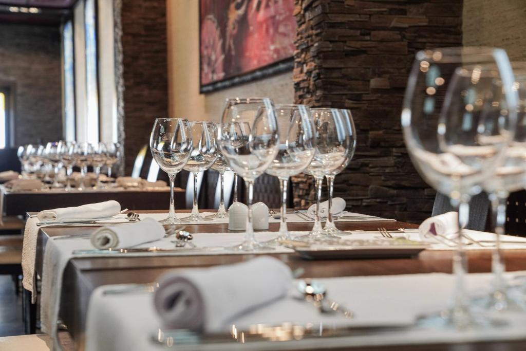 Eleganti sakārtota galda iestatījums restorānā ar glītām glāzēm un salvetēm, kas rada svinīgu atmosfēru. Interjerā redzami akmens elementi un lieli logi, kas ļauj ieplūst dabiskajai gaismai.