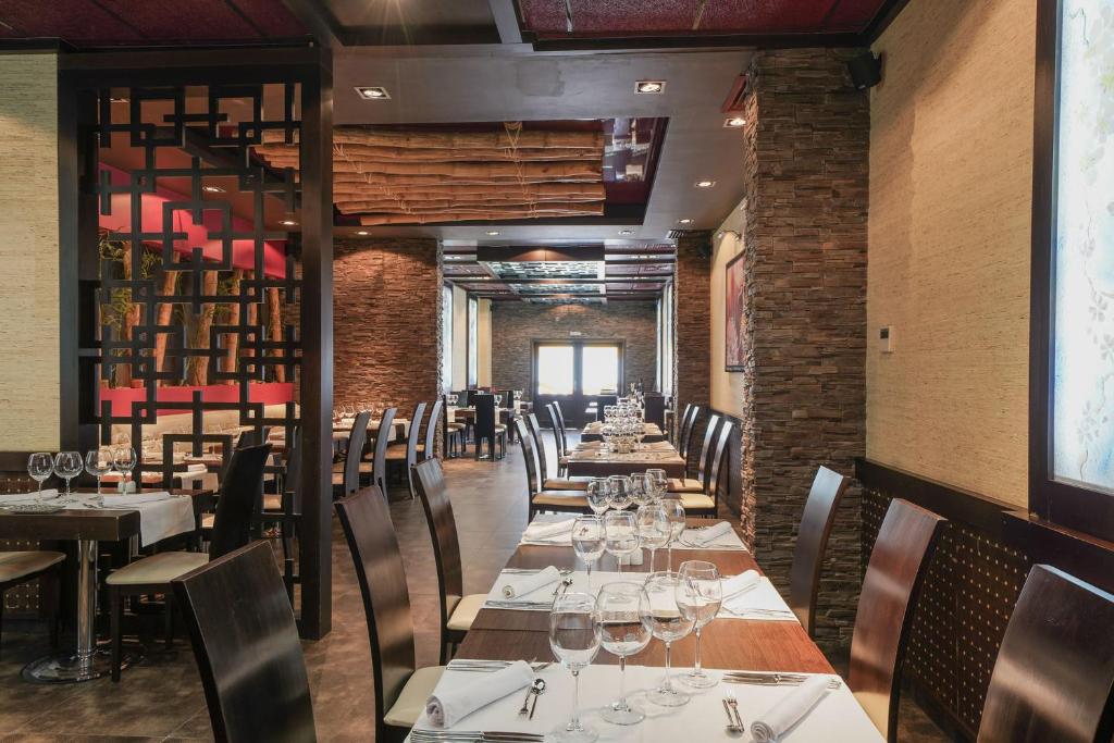 Mūsdienīgs restorāns ar elegantu interjeru, kas ietver koka un akmens elementus, ar galdiem, kas sakārtoti rindu. Svaigi klāti galdi ar glāzēm un galda piederumiem, radot patīkamu atmosfēru.