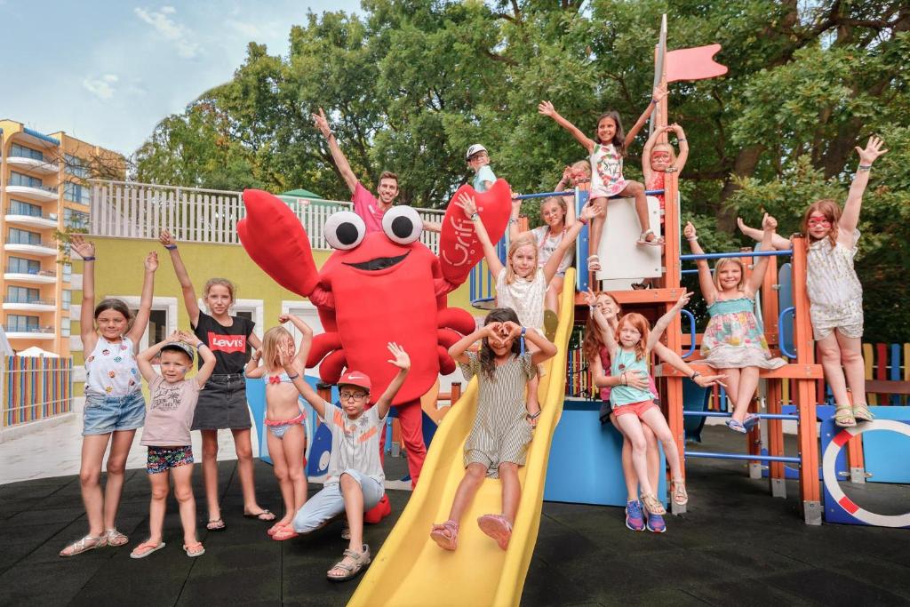 Bērni un pieaugušais jautrā atmosfērā spēļu laukumā, kur viņi pozē kopā ar lielu krabja maskotu. Krāsains fons un priecīgas sejas rada jautru un draudzīgu noskaņu.