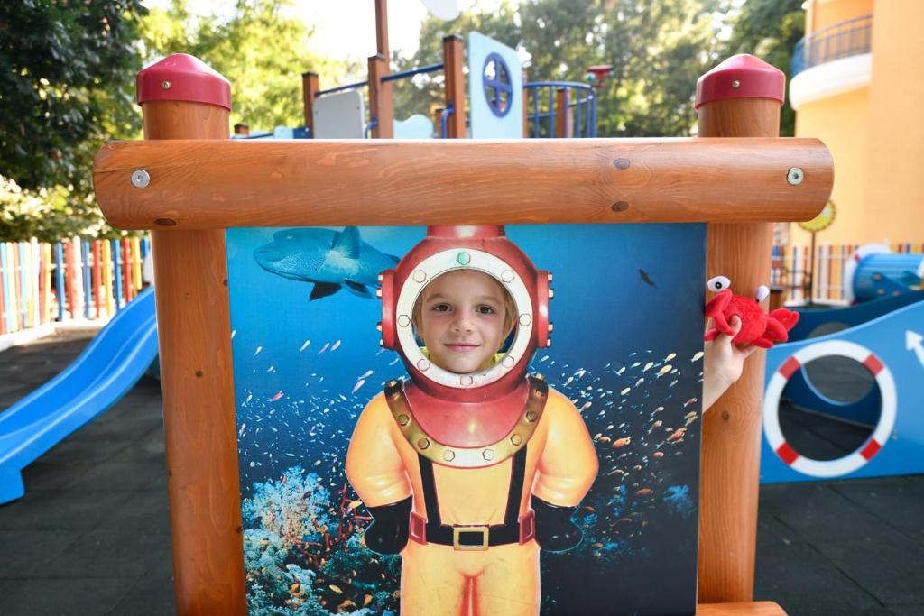Bērns izskatās kā zemūdens pētnieks, skatoties no spēļu laukuma paneļa ar jūras ainavu fonā. Spēļu laukums ir krāsains un piedāvā dažādas aktivitātes.