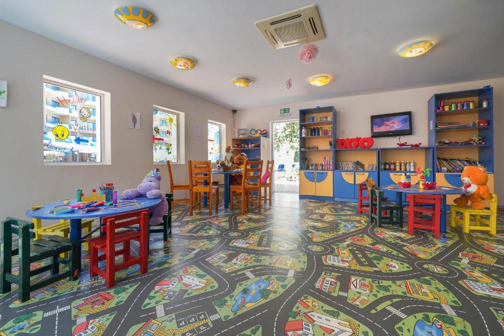 Krāsaina bērnu rotaļu telpa ar spēļu galdiem un rotaļlietām, kas izkārtotas uz ceļa raksta grīdas. Telpā ir logi ar zīmējumiem un plaukti ar mākslinieciskiem materiāliem.