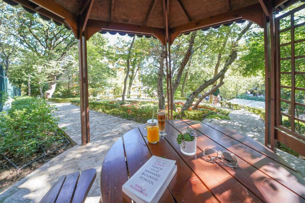 Mierīga ainava ar koka paviljonu, kur uz galda atrodas grāmata, dzēriens un puķu pods, apkārt zaļojoši koki un ceļi. Ideāla vieta relaksācijai un lasīšanai.