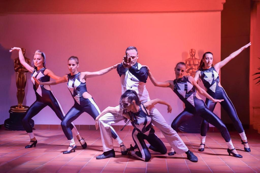 Deju grupa izpilda dinamisku priekšnesumu, pārvietojoties ritmā un izceļot stilizētas kostīmu formas. Fons ir apgaismots ar siltām krāsām, radot enerģisku atmosfēru.